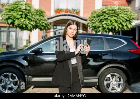 Junge, ernsthafte, dunkelhaarige Geschäftsfrau mit Tablet im Freien, die vor der neuen schwarzen Autoüberkreuzung und dem modernen Backstein-Ofiice-Gebäude steht Stockfoto