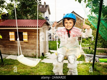 Kleiner Junge aus supersicherem Sprudelmaterial und überschützende Mutter schwingen auf Schaukeln im Garten Stockfoto