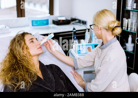 Weiblicher Arzt und Patient während der Untersuchung der Gesichtshaut. Die Ergebnisse des Hautzustands werden auf dem Display angezeigt. Stockfoto