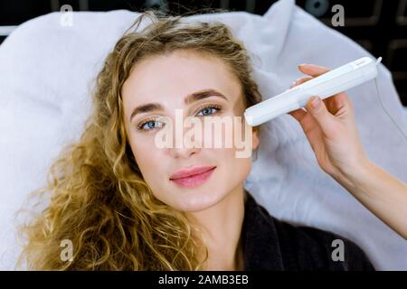 Nahaufnahme der Hand des Arztes, der Anti-Aging-Verfahren in der Kosmetologie-Praxis macht. Zufriedene junge Frau auf der Couch liegend, lächelnd und entspannend Stockfoto