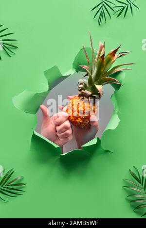 Kleine reife orangefarbene Ananas in menschlicher Hand, die andere Hand zeigt ein OK-Zeichen. Hände mit dem Obst zeigen aus dem gerippten Papierloch. Tropisch-grüne Geometrie Stockfoto