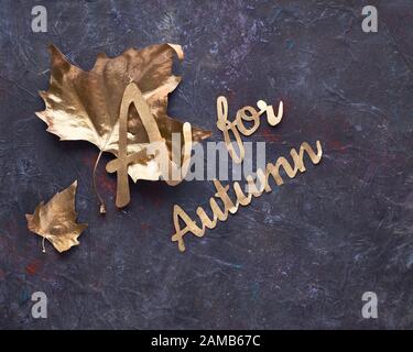 Herbst, kreatives Flachlay. Draufsicht auf die goldfarbenen, dunkelgrauen, strukturierten Blätter. Text "A oder Herbst" in vergoldetem Italic Scrip Stockfoto