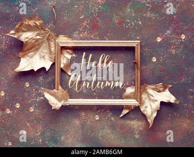 Flache Herbstlage mit vergoldeten Lamellen und vergoldetem Text "Hello Autumn" in goldenem Rahmen auf dunkelbrauner strukturierter Leinwand. Draufsicht, dekorative Zierkarre Stockfoto