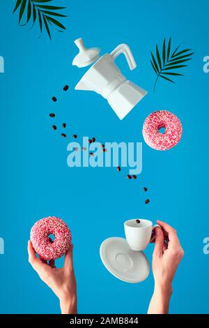 Trendy Levitation. Fliegen Linie der Kaffeebohnen zwischen Keramik Kaffeemaschine und Espresso Tasse mit Untertasse. Zwei weibliche Hände halten rosa Donuts mit su Stockfoto