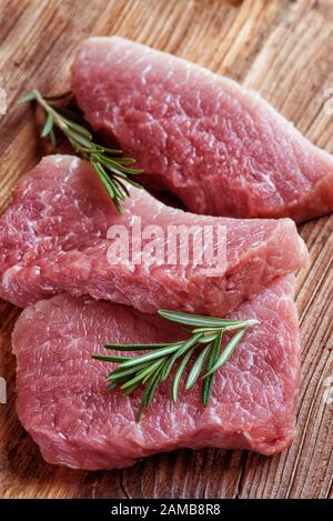 Drei Steaks aus frischem Bio-Fleisch und zwei Rosmarinsprengstücke auf einer alten Holztafel liegen dicht beieinander Stockfoto