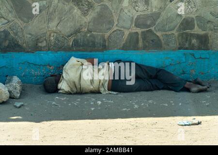 Ein obdachloser kenianischer Mann, der im Schatten eines Gebäudes auf einem sandigen und staubigen Straßenbelag schläft, Mombasa, Kenia Stockfoto