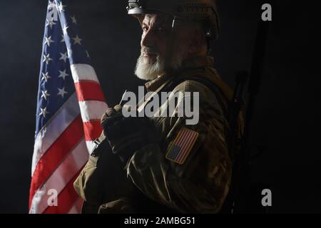 Seitenansicht des alten Militäroffiziers mit Blick auf die Kamera. Ernte eines bärtigen amerikanischen Veteranen in Uniform und Helm, der mit einer Pistole auf der Schulter auf schwarzem Hintergrund mit Fahne posiert. Begriff des Militärs, der Armee. Stockfoto