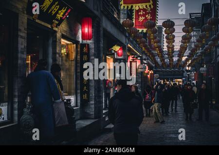 Menschen, die in Pekings Hutong spazieren und einkaufen. Einige Männer tragen ein traditionelles chinesisches Kleid namens Changshan. Über einer typischen chinesischen Lampe. Stockfoto