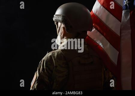 Rückansicht des inkogniito alten Militärbeamten, der die Fahne hält und wegschaut. Nahaufnahme des bärtigen amerikanischen Veteranen in Uniform und Helm auf schwarzem Hintergrund. Begriff des Militärs, der Armee. Stockfoto