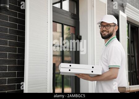 Seitenansicht des fröhlichen jungen männlichen Kuriers in weißem Polo und Kappe mit Pizza im Freien. Bärtiger Mann in einer Brille, die in der Nähe der Tür steht, zwei Pizzakisten hält und lächelt. Konzept für die Lieferung von Lebensmitteln. Stockfoto