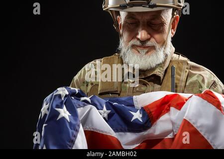 Ernte von bärtigen amerikanischen Militärveteranen, die Flagge in Waffen halten. Porträt des alten stolzen männlichen Beamten in Tarnuniform und Helm in dunkler Atmosphäre. Begriff des Militärs, Patriotismus. Stockfoto