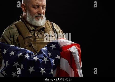 Ernte von bärtigen amerikanischen Militärveteranen, die Flagge in Waffen halten. Porträt des alten stolzen männlichen Beamten in Tarnuniform, der in dunkler Atmosphäre posiert. Begriff des Militärs, Patriotismus. Stockfoto