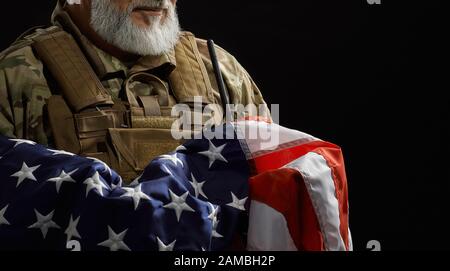 Nahaufnahme des bärtigen amerikanischen Militärveteranen, der die Flagge in Waffen hält. Porträt von inkognito altem stolzen männlichen Offizier in Tarnuniform, der in dunkler Atmosphäre posiert. Begriff des Militärs, Patriotismus. Stockfoto