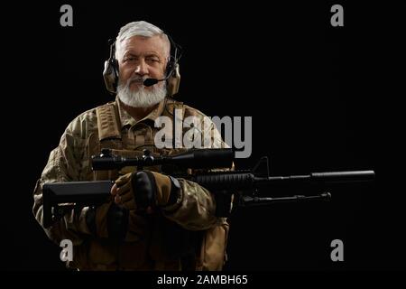 Vorderansicht des alten Militärbeamten mit Gehörschutz, der das Scharfschützengewehr hält und die Kamera betrachtet. Porträt des bärtigen Veteranen in Uniform posiert mit Waffe auf schwarzem Studiohintergrund. Stockfoto