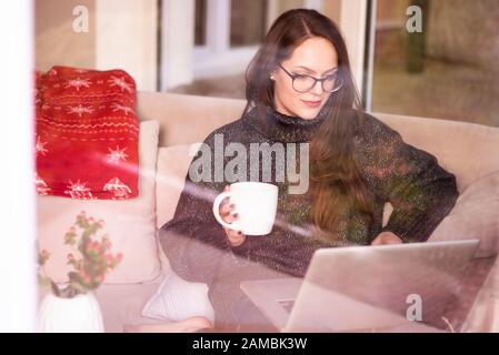 Schuss von junger Frau, die auf Laptop arbeitet und Tee trinkt, während sie zu Hause hinter dem Glasfenster auf dem Sofa sitzt. Heimbüro. Stockfoto