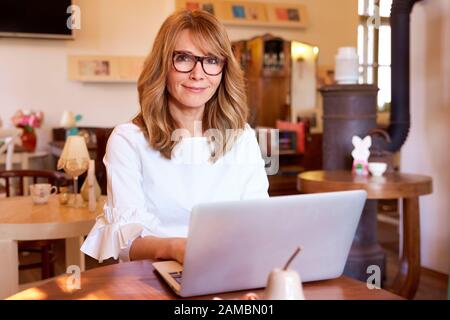 Schuss von Frau mittleren Alters, die am Schreibtisch sitzt und in einem kleinen Café an einem Notizbuch arbeitet. Kleine Unternehmen. Stockfoto