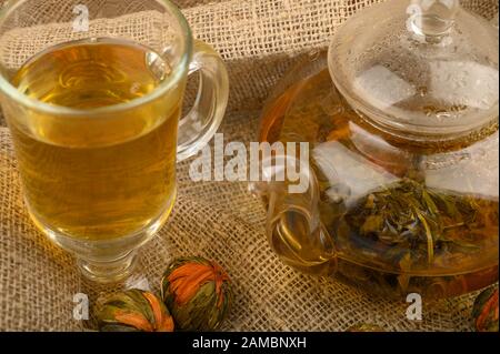 Blumentee, der in einer Glaskanne gebrüht wird, ein Glas Tee und Blumenkugeln auf dem Hintergrund rauer Homespun-Qualität. Nahaufnahme Stockfoto