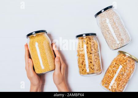 Die Hände der Frau, die Glasbecher aus Hirse neben anderen Groats und Pasta in umweltfreundlichen Aufbewahrungsbehältern halten. Stockfoto