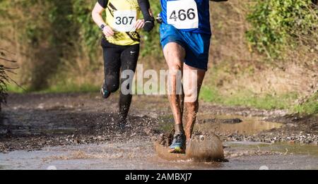 Beine und Füße von zwei weißen männlichen Läufern, die sich darüber tupften Eine schlammige Pfütze in einem Winterhalbmarathon-Langlauf-Rennen Tragen Lätzchen mit Rennnummern Stockfoto