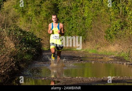 Entschlossener weißer Mann, weiße Frau, die an einem sonnigen Januartag durch eine große schlammige Pfütze in einem Winterhalbmarathon auf einer Landstraße läuft Stockfoto