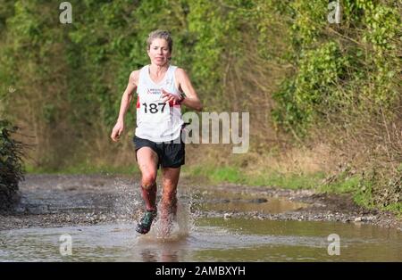 Entschlossener weißer Mann, weiße Frau, die an einem sonnigen Januartag durch eine große schlammige Pfütze in einem Winterhalbmarathon auf einer Landstraße läuft Stockfoto