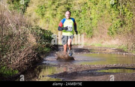 Entschlossener weißer Mann, weiße Frau, die an einem sonnigen Januartag durch eine große schlammige Pfütze in einem Winterhalbmarathon auf einer Landstraße läuft Stockfoto