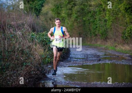 Entschlossener weißer Mann, weiße Frau, die an einem sonnigen Januartag durch eine große schlammige Pfütze in einem Winterhalbmarathon auf einer Landstraße läuft Stockfoto