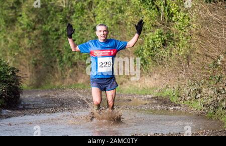 Entschlossener weißer Mann, weiße Frau, die an einem sonnigen Januartag durch eine große schlammige Pfütze in einem Winterhalbmarathon auf einer Landstraße läuft Stockfoto