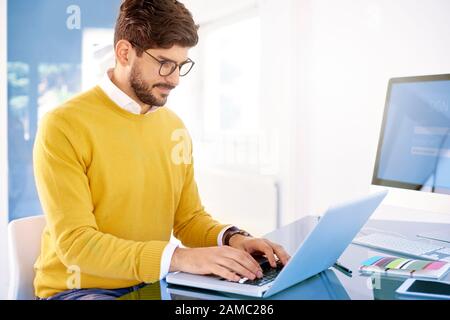 Portrait der junge Geschäftsmann mit seinem Laptop und etwas eingeben, während im Büro sitzen und arbeiten. Stockfoto