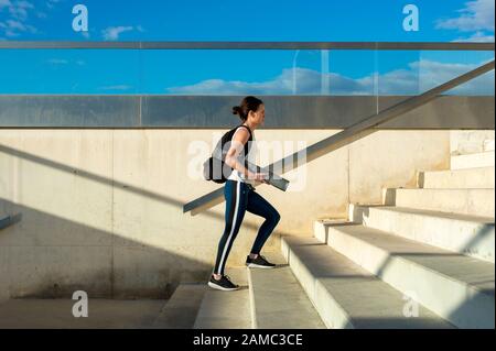 Sportliche Frau, die auf Stufen geht und eine Trainingsmatte und einen Rucksack mit Sportbekleidung trägt. Stockfoto