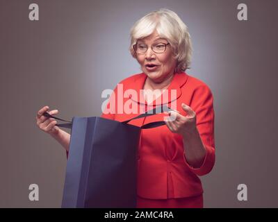 Überraschte Seniorin, die in Papiertasche, Verkauf oder Geschenkkonzept schaut Stockfoto