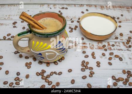 Mexikanischer Kaffee mit Zimt in Tasse mit mexikanischer Dekoration auf weißem Holztisch mit organischen Kaffeebohnen und natürlicher Schüssel mit Milch und Holzspoo Stockfoto