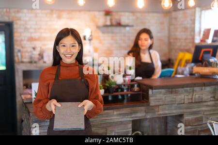 Junge asiatische weibliche Kellnerin trägt Schürze und zeigt dem Geschäftsinhaber Essen und Getränke Café-Konzept. Stockfoto