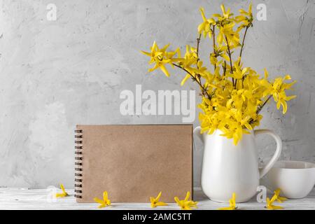 Stillleben mit schönen frühlingsgelben Forsythia Blumen, Kaffeetasse und leerer Grußkarte auf konkretem Hintergrund. Verspotten Sie sich. Urlaub oder Hochzeit Co Stockfoto