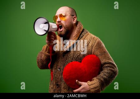 Schreiender Mann in rosafarbener Brille hält Spielzeugherz und schreit, während er im Studio auf leerem grünem Hintergrund steht Stockfoto