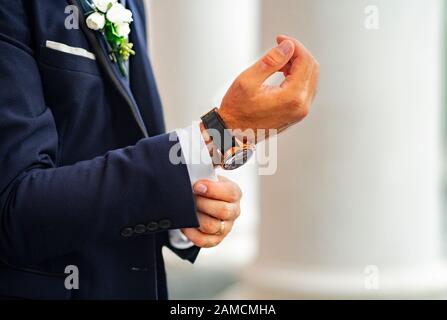 Der Bräutigam in einem teuren, klassischen blauen Anzug mit einem Blumenstrauß am Revers passt den Manschettenknauf an der Hemdmanschette an. Hände eines Mannes in einem Anzug mit einem Expen Stockfoto