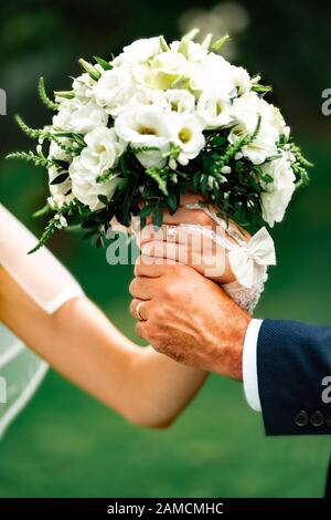 Die Hände der Frau und des Bräutigams verflochten sich und halten einen Hochzeitsstrauß zart weißer Rosen, Eustomas. Nahaufnahme. Hände der Neuvermählten in der Hochzeit rin Stockfoto