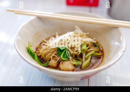 Nahaufnahme der Würzigen thailändischen Bootsreis-Noodle-Suppe Kuai Tiao Ruea, auf einer Keramikschale auf weißem Holztisch servieren. Stockfoto