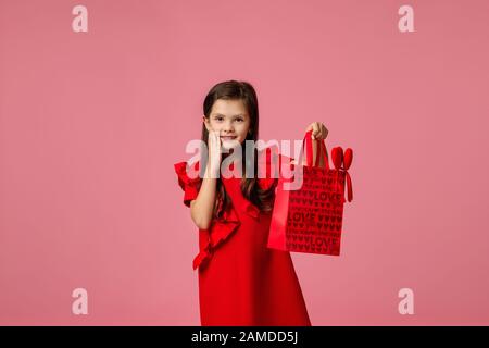 Valentinstag. Lächelndes Kindermädchen, das rote Papier-Einkaufstasche auf pinkfarbenem Hintergrund hält. Shopping-Verkauf. Copy-Platz Stockfoto