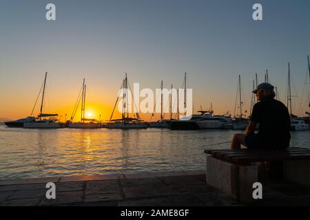 Naxos Griechenland - 11. August 2019; nam fängt Sonne auf der Seite des Gesichts in Silhouette sitzend und bewundert den Sonnenaufgang und die Yacht-Masten, die alle von goldener Stunde beleuchtet werden Stockfoto