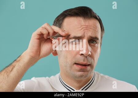 Kopf und Schultern Portrait des winkenden Mannes mittleren Alters, der Augenbrauen zupft, die vor blauem Hintergrund stehen Stockfoto