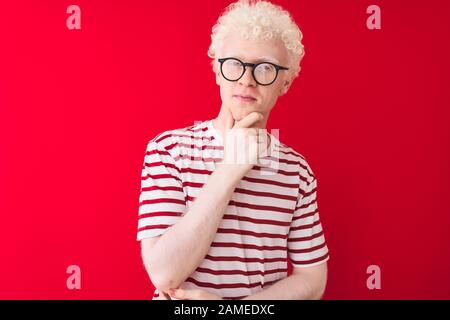 Junger albino blonder Mann mit gestreiftem T-Shirt und Brille über isoliertem rotem Hintergrund, der mit einem Lächeln mit gekreuzten Armen a zuversichtlich auf die Kamera blickt Stockfoto
