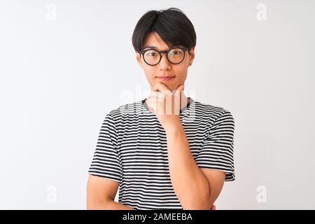 Der Chinese, der eine Brille trägt, und das marineblau gestreifte T-Shirt, das über einem isolierten weißen Hintergrund steht und mit einem Lächeln und einem gekreuzten A sicher auf die Kamera blickt Stockfoto