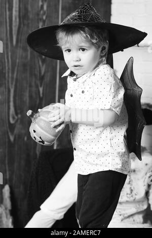 Junge auf Holz und weißem Hintergrund. Kind mit verängstigte Gesichter in Hexenhüten spielt mit geschnitzten Kürbissen. Halloween- und Urlaubskonzept. Der kleine Zauberer macht Halloween-Dekor. Stockfoto