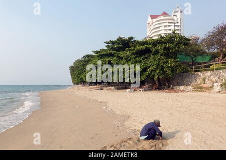 Pattaya, Thailand - 23. Dezember 2019: Ältere Frau sitzt am Strand und versucht, Muscheln zu finden. Ausländertouristen in Sonnenliegen im Hintergrund. Stockfoto