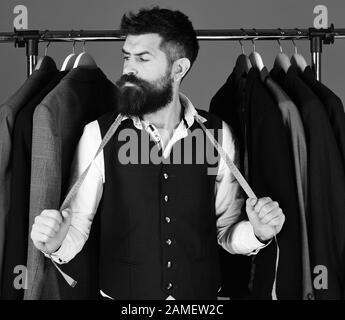 Mann mit Bart am Kleiderständer. Mit ernsthaftem Gesicht und Messband in der Nähe von individuellen Jacken auf blauem Hintergrund zuschneiden. Mode- und Nähkonzept. Der Designer ergreift Maßnahmen in der Nähe von Kleiderbügeln. Stockfoto
