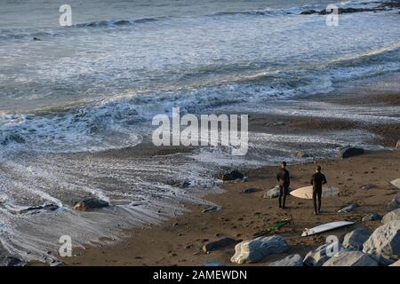 Aberystwyth Ceredigion UK 10. Januar 2019: Zwei Surfer mit Surfbrettern am Strand bereiten sich darauf vor, ihren Freund bereits im Meer zu begleiten Stockfoto