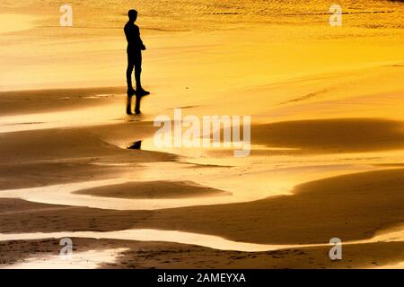 Eine einsame männliche Figur, die von dem intensiven Licht der untergehenden Sonne umschimmert wird, während er sich am Strand von Fistral in Newquay in Cornwall auf das Meer blickt. Stockfoto