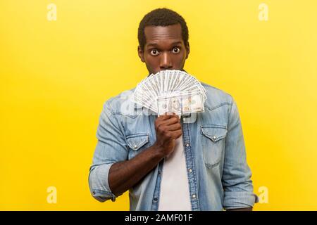 Portrait des überraschten verängstigte Mannes in einem Jeanshemd, das Dollarscheine hält, während er aus dem Geld schaufelt, während er das halbe Gesicht mit staunenden schockierten Augen versteckt. Stockfoto