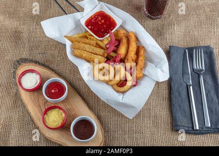 Imbissplatte - leckerer knolliger Zwiebelring, pommes frites, Wurst, gebratener Käse, heißer Imbiss. Spezielles Gewürz und Koks auf Leinengewebe. Stockfoto